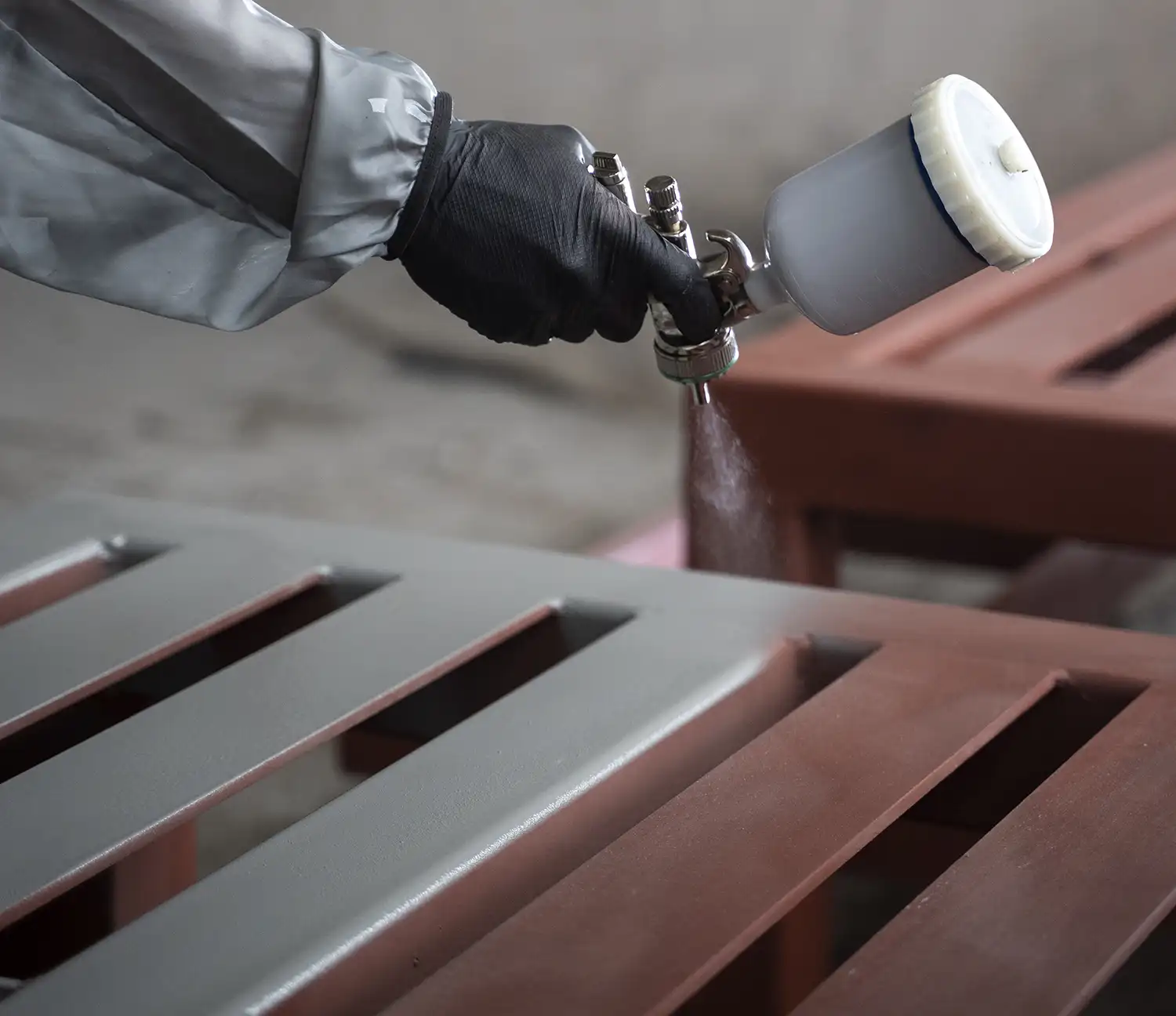 Mano con guante negro y pistola de pintura aplicando pintura gris sobre una superficie metálica con rejillas. Proceso de pintado industrial.