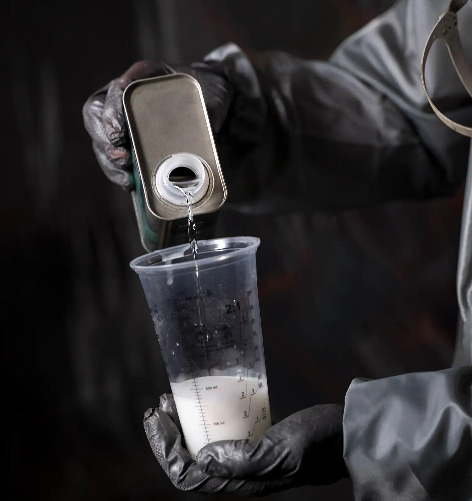 Persona con guantes de seguridad vertiendo líquido transparente de una lata metálica en un vaso medidor con líquido blanco, preparando una mezcla industrial.